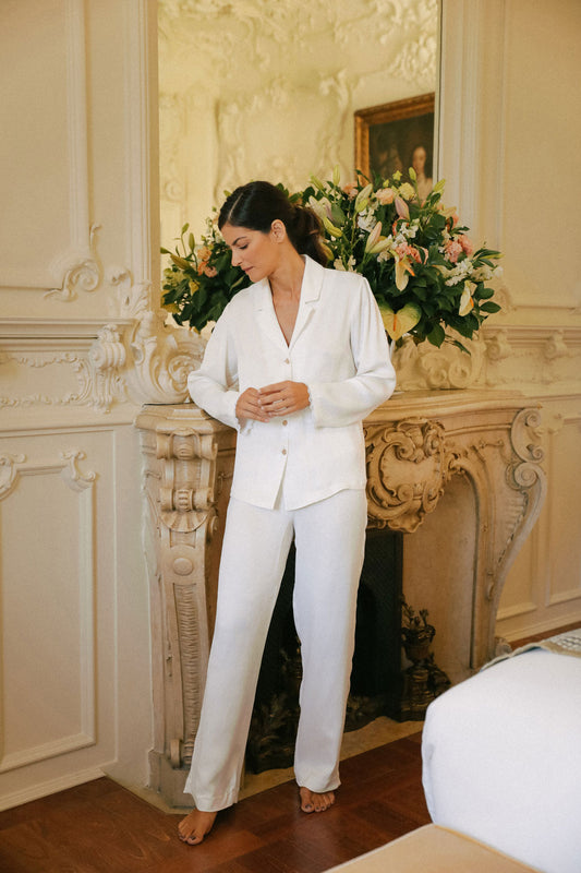 woman standing, leaning against a large fireplace, dressed in a white silk pajama set.