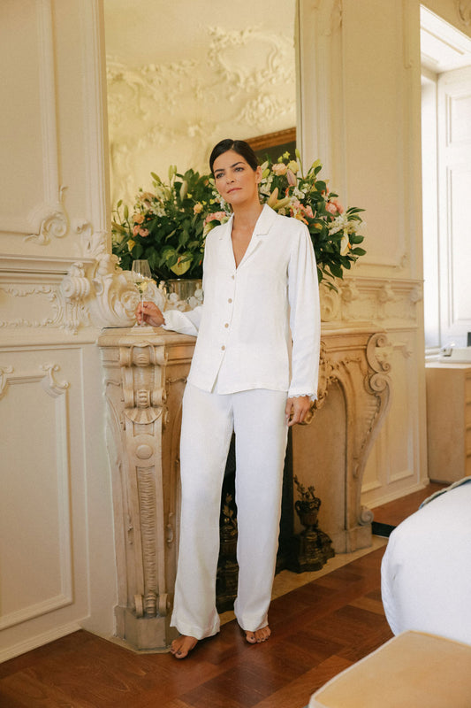woman standing, leaning against a large fireplace, dressed in a white silk pajama set.